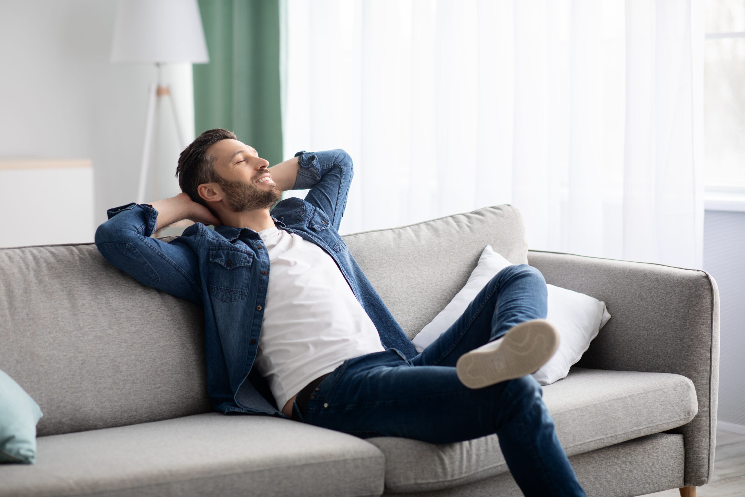 Happy bearded man in casual clothes relaxing on couch at home, looking at copy space, side view. Relaxed middle-aged man having break on sofa in living room, dreaming about prosperous life